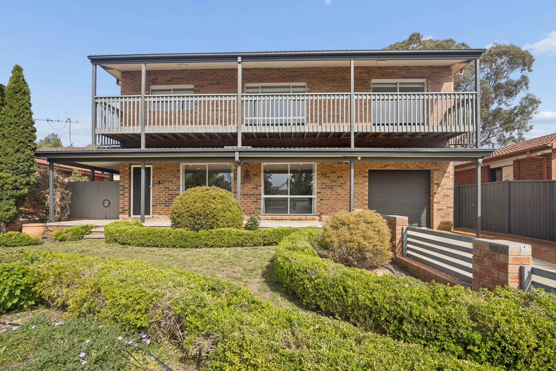 Two-storey brick house with a full-width upstairs balcony, hedges and lawn